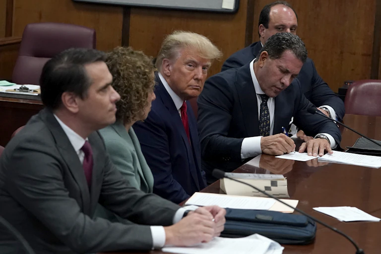 A white man with yellow hair and a suit sits at a table with other people in formal clothing him around him and looks toward the camera with a serious face.