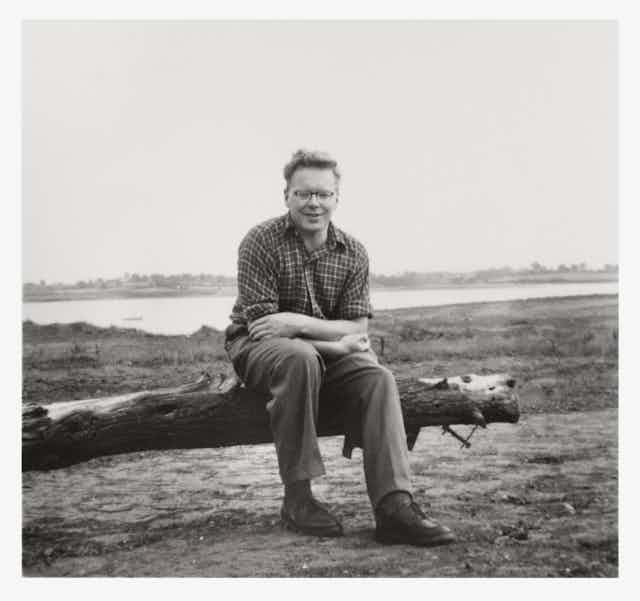 Black and white photo, man in checked shirt and trousers sat on fallen branch of tree, estuary landscape in background