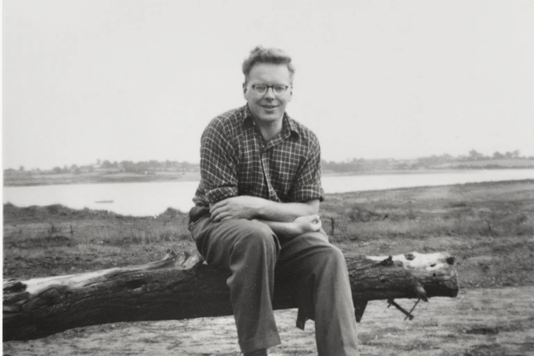 Black and white photo, man in checked shirt and trousers sat on fallen branch of tree, estuary landscape in background