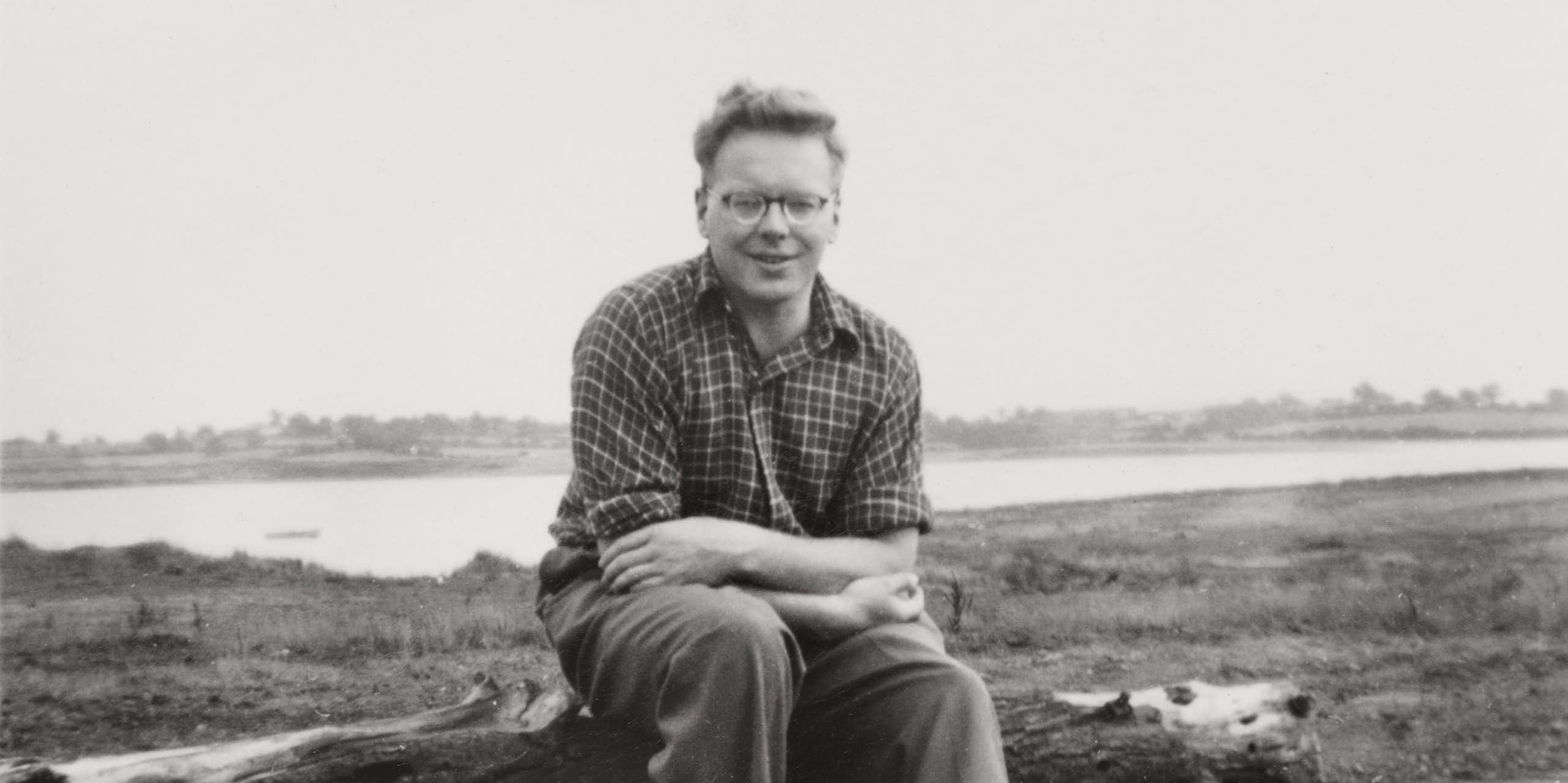 Black and white photo, man in checked shirt and trousers sat on fallen branch of tree, estuary landscape in background