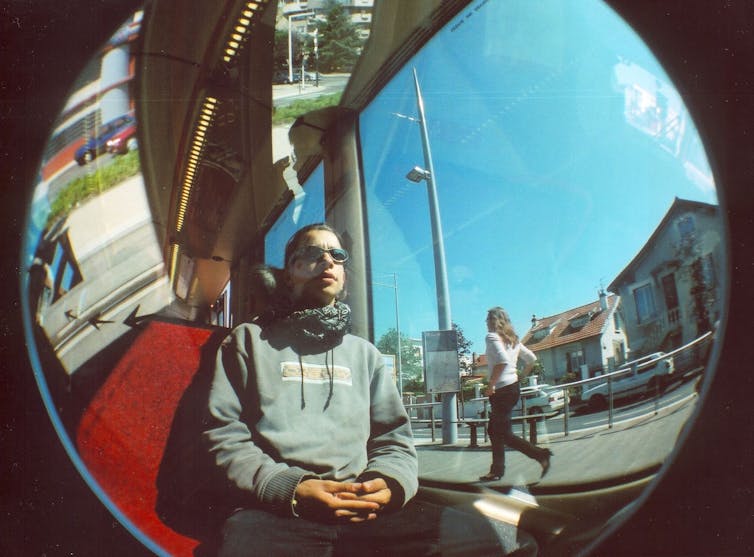 Young man seated in a tram with woman walking past outside, captured by a fisheye camera lens