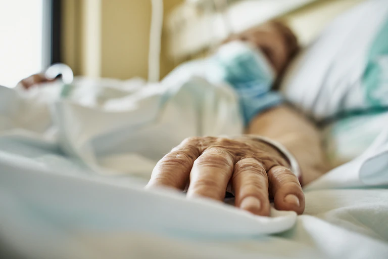 Patient wearing mask lying on hospital bed, hand extended toward camera