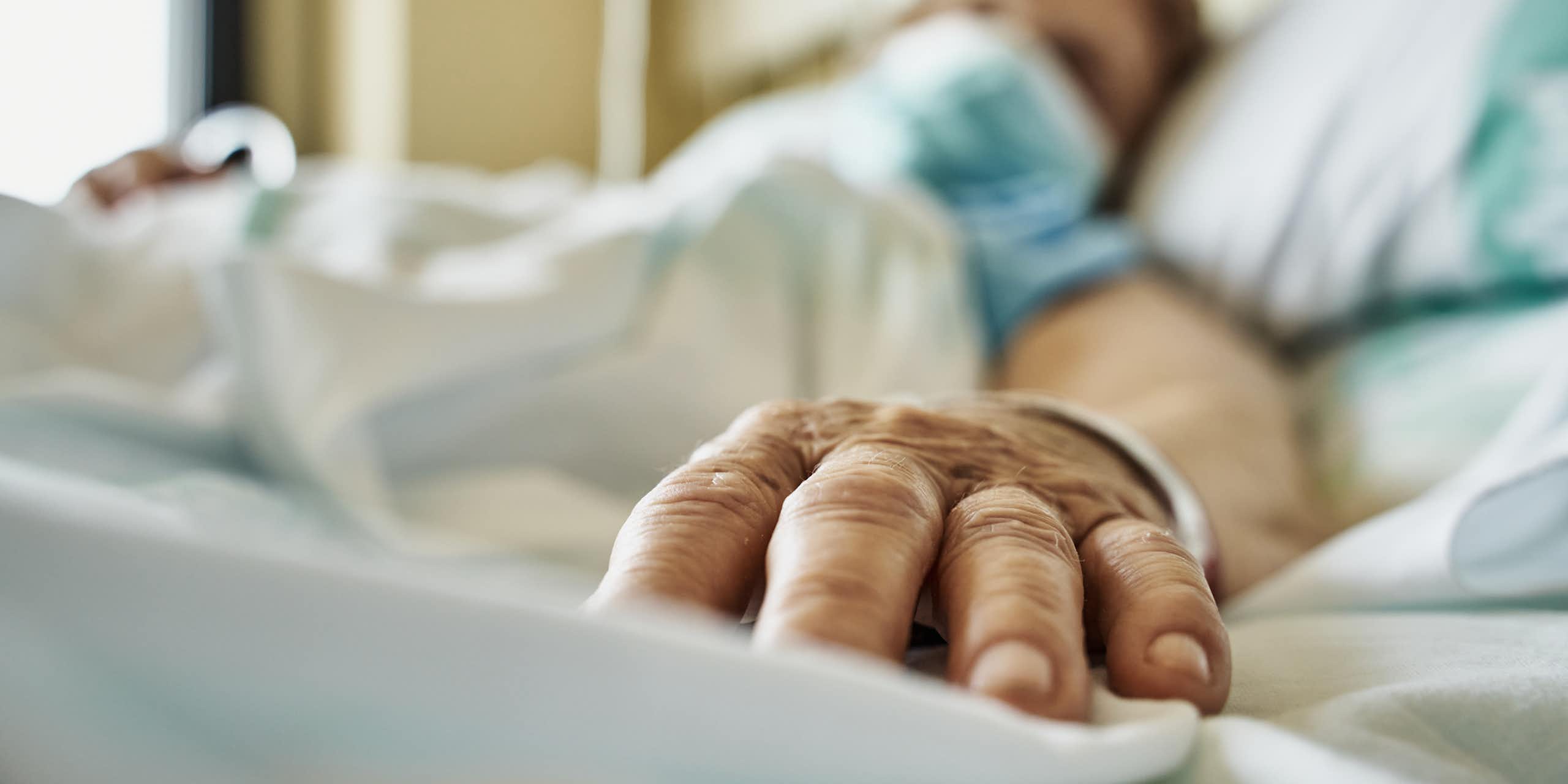 Patient wearing mask lying on hospital bed, hand extended toward camera