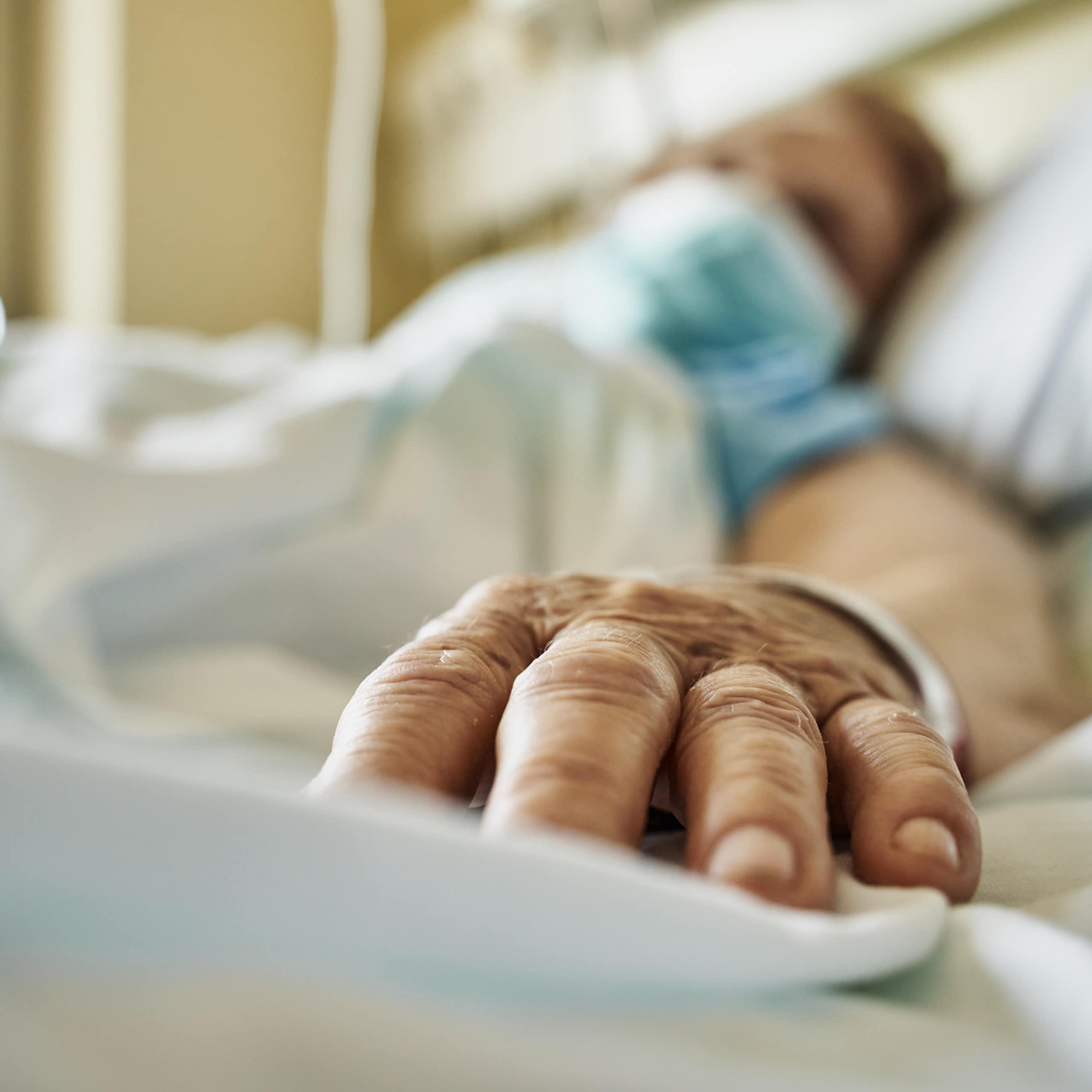 Patient wearing mask lying on hospital bed, hand extended toward camera