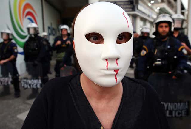 A protester standing in front of police, wearing a white mask with fake blood coming from its nose and mouth