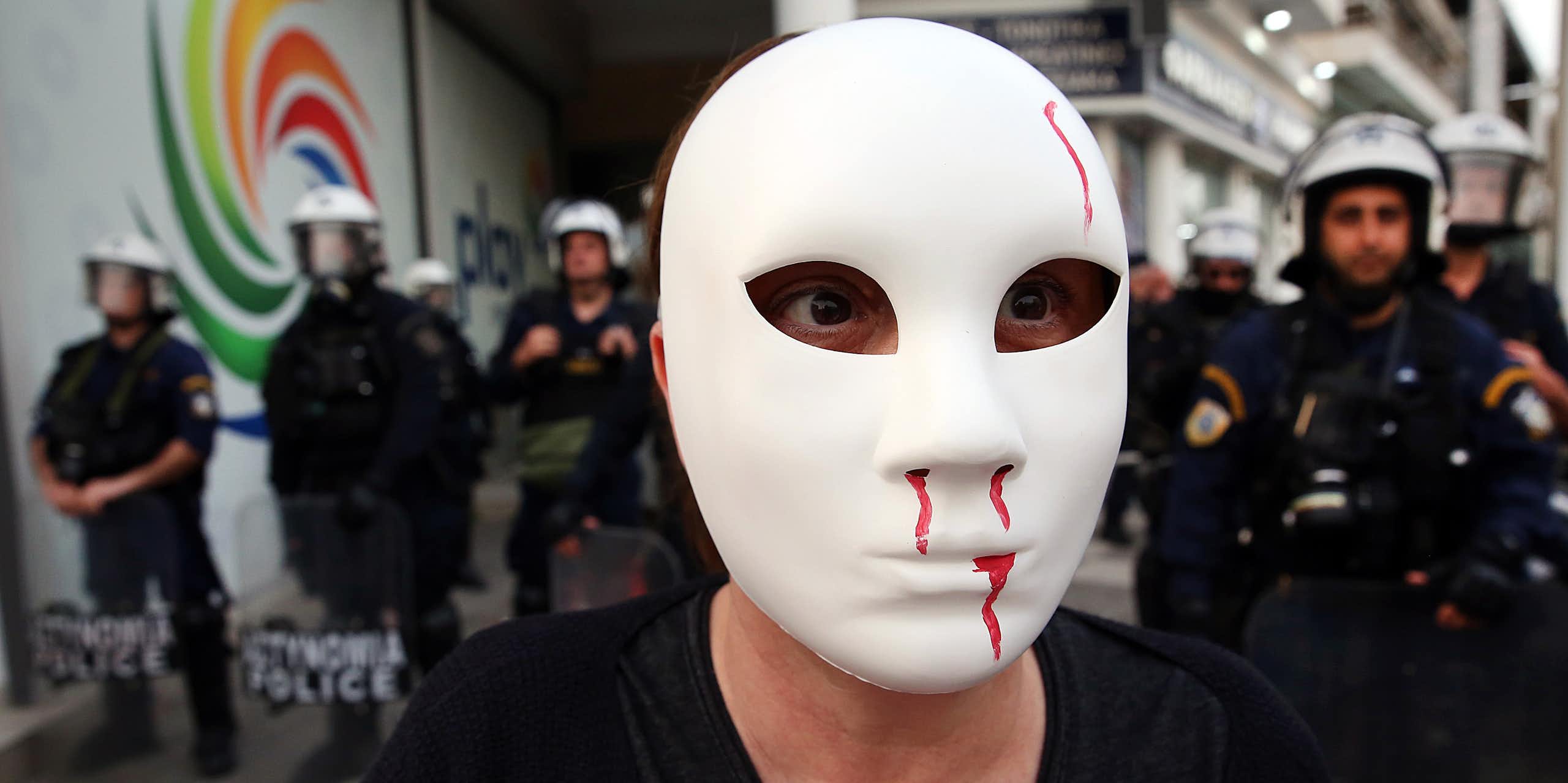 A protester standing in front of police, wearing a white mask with fake blood coming from its nose and mouth