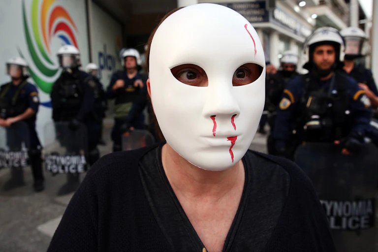 A protester standing in front of police, wearing a white mask with fake blood coming from its nose and mouth