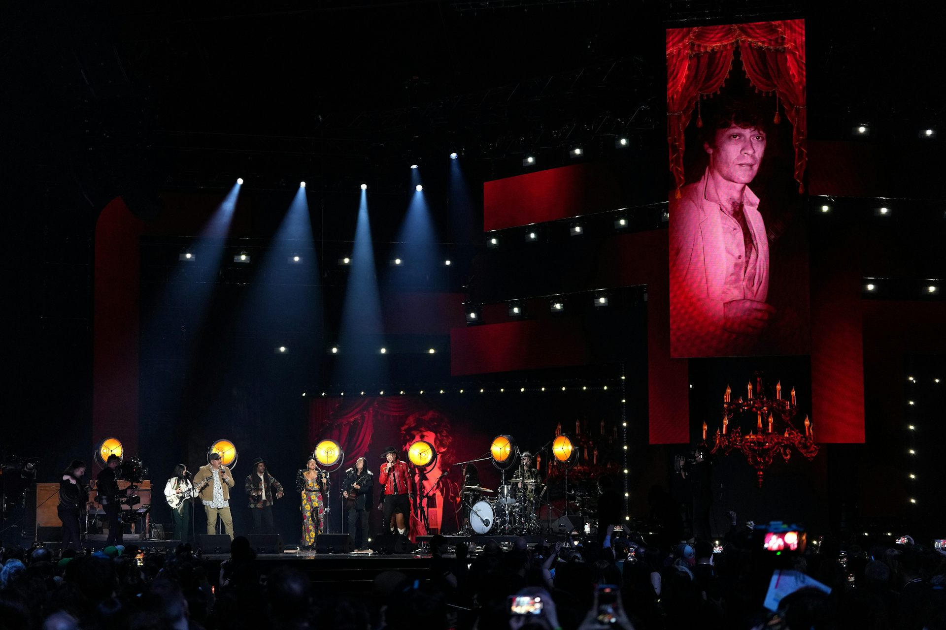 Performers seen on a stage with the image of a person projected on a screen behind them.