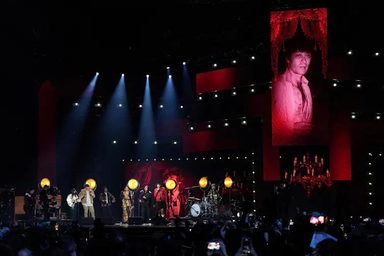 Performers seen on a stage with the image of a person projected on a screen behind them.