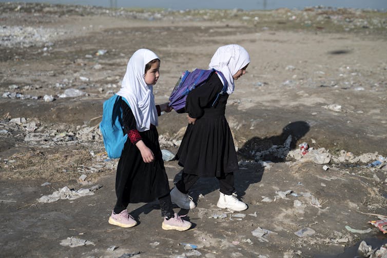Two girls wearing white headscarves, sneakers, long black overgarments and carrying colorful backpacks, walk on a rocky, trash-strewn tract.