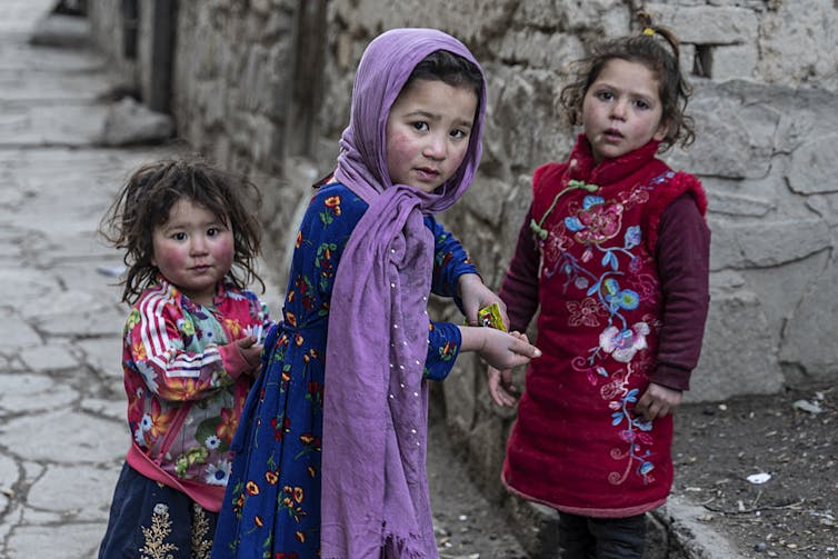 Tres niñas con prendas coloridas comen bocadillos en una calle.