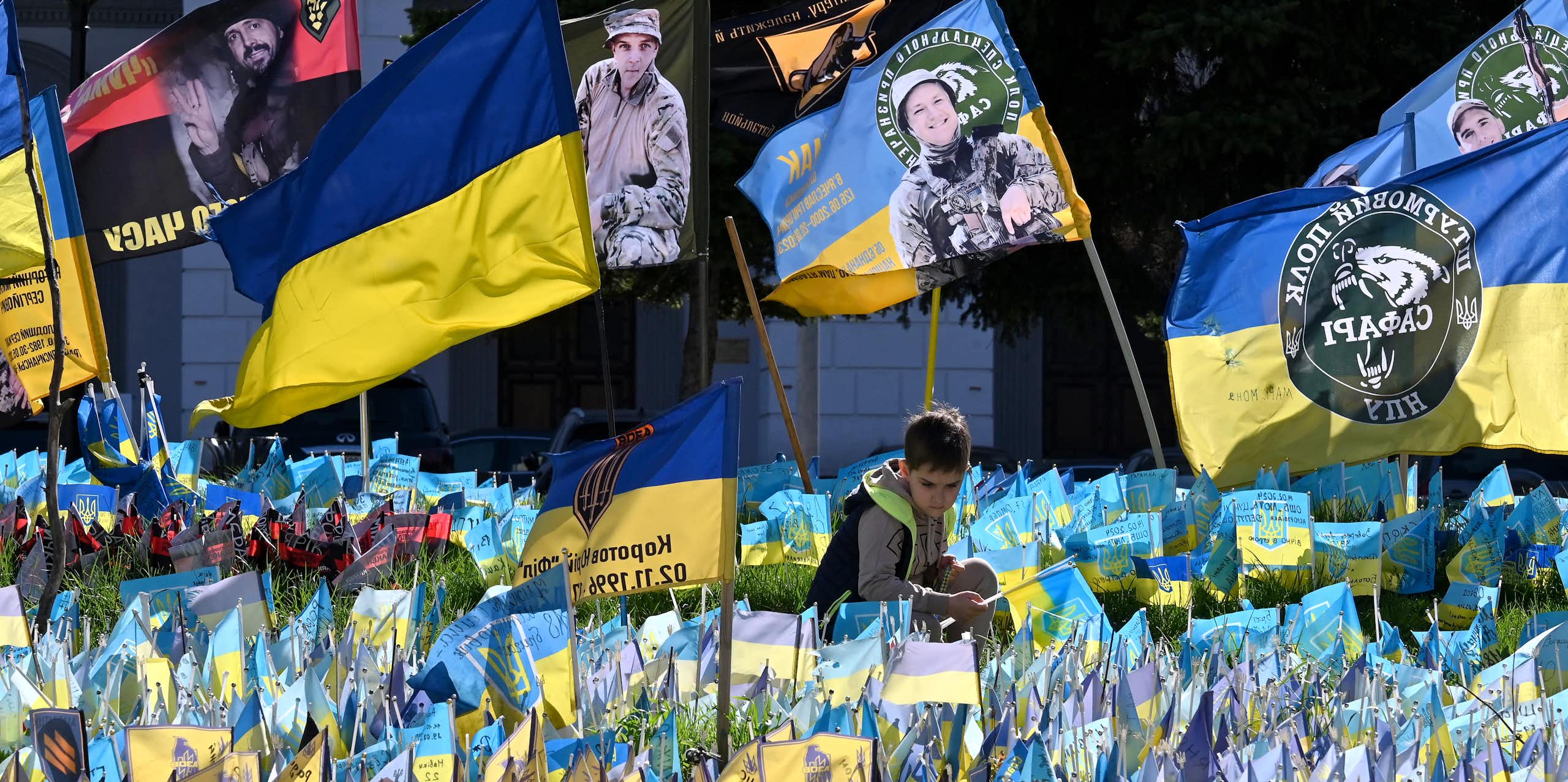 A boy sits among many yellow and blue flags, including some with photos of soldiers and Ukrainian writing.
