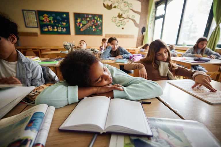 Elementary aged child has head down on desk with other kids sitting at desks in a classroom.