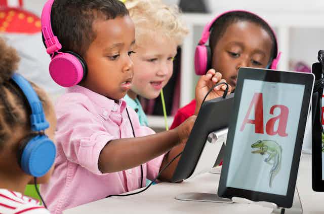 Four small kids wearing bright colored headphones look at digital tablets on their desks.