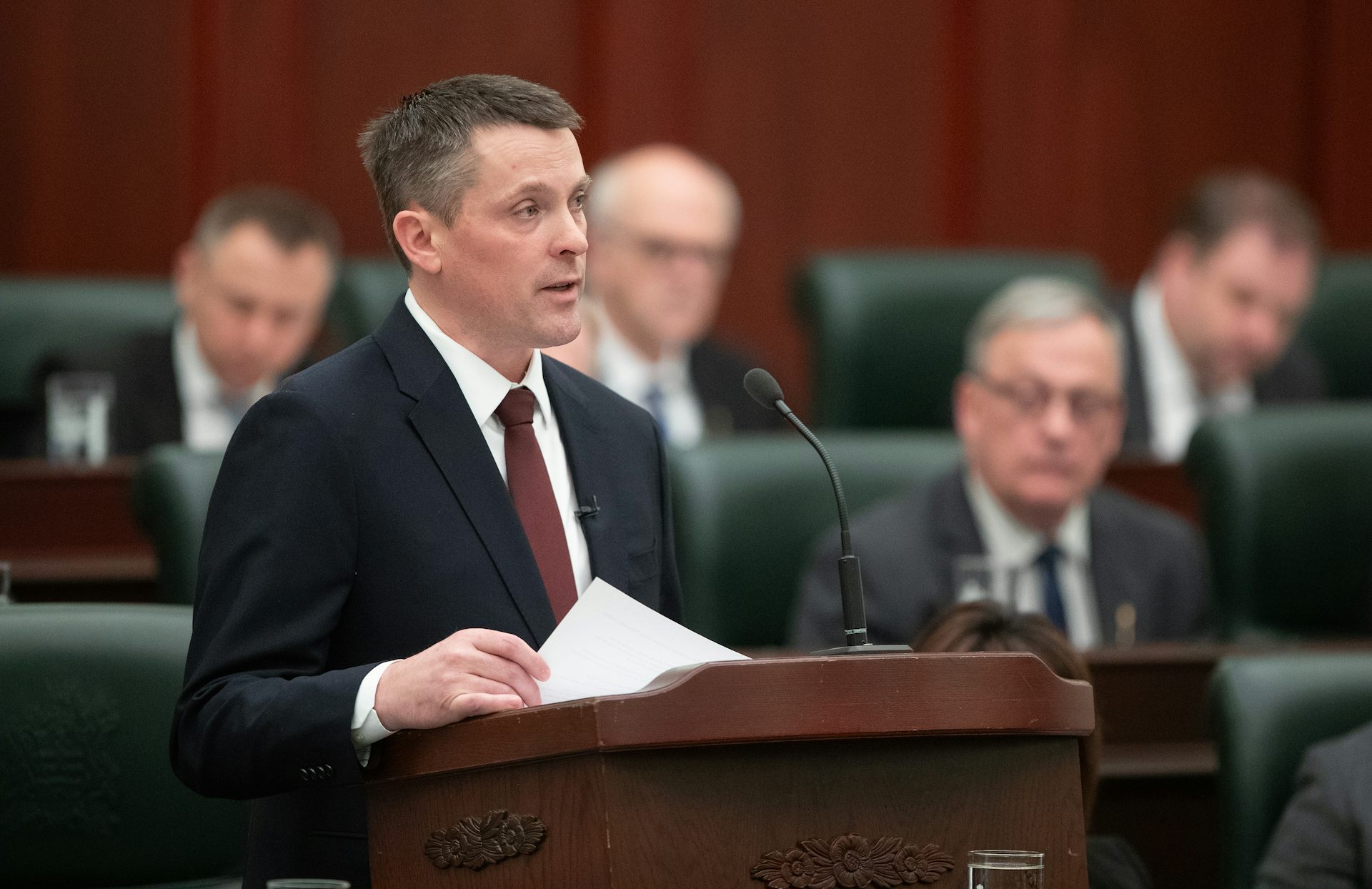 A middle-aged white man in a suit speaks into a microphone from behind a podium