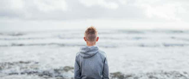 Young person in a hoodie looking out across the ocean, with back to camera