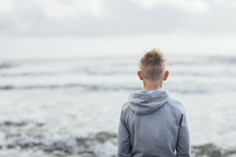 Young person in a hoodie looking out across the ocean, with back to camera