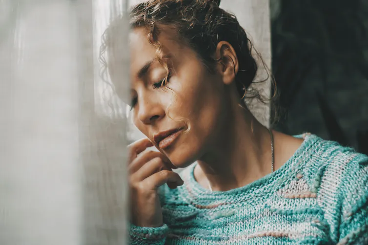 A tired woman leaning her head on a window with her eyes closed.
