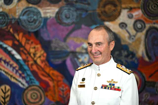 A man in a white navy uniform smiles in front of a multicoloured backdrop