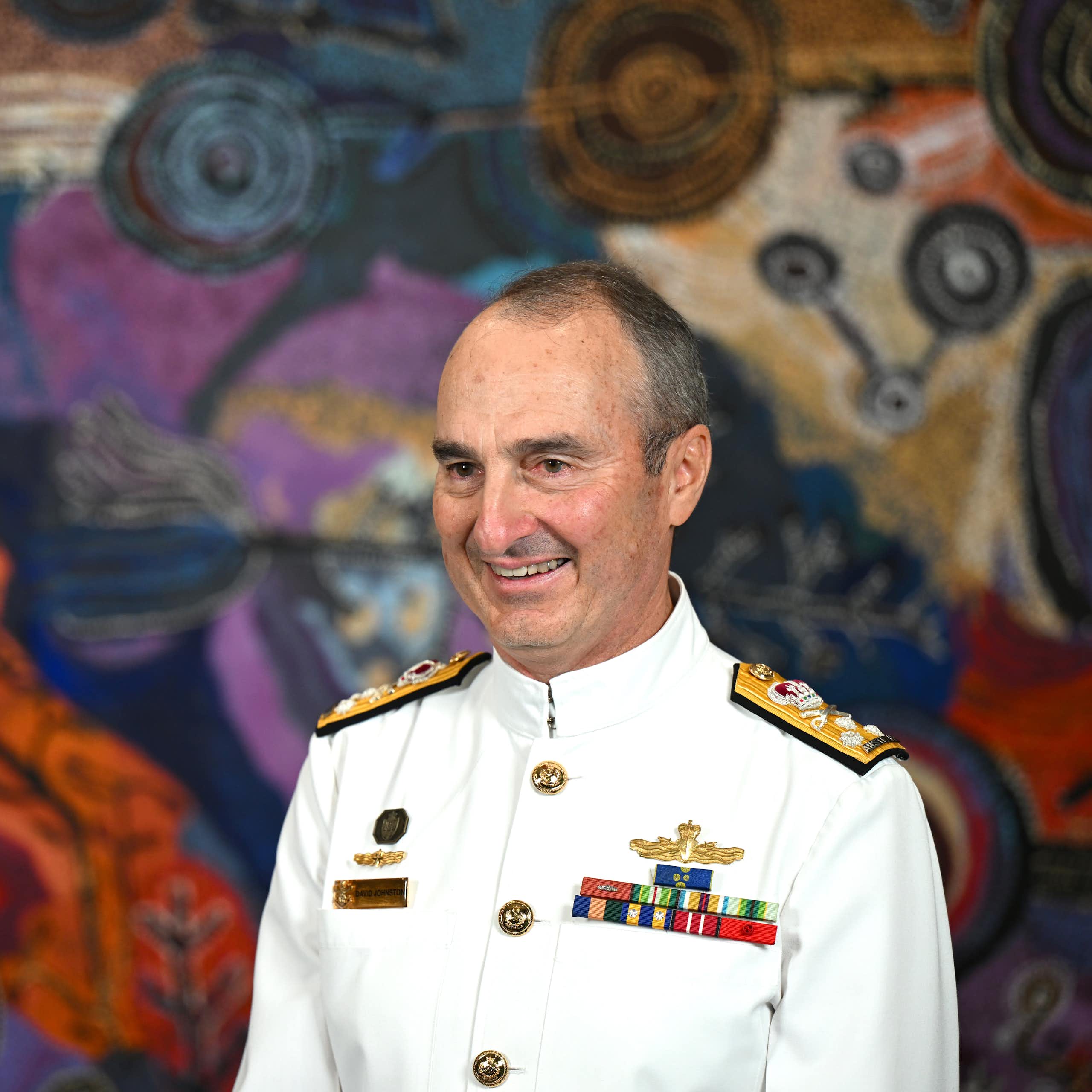 A man in a white navy uniform smiles in front of a multicoloured backdrop
