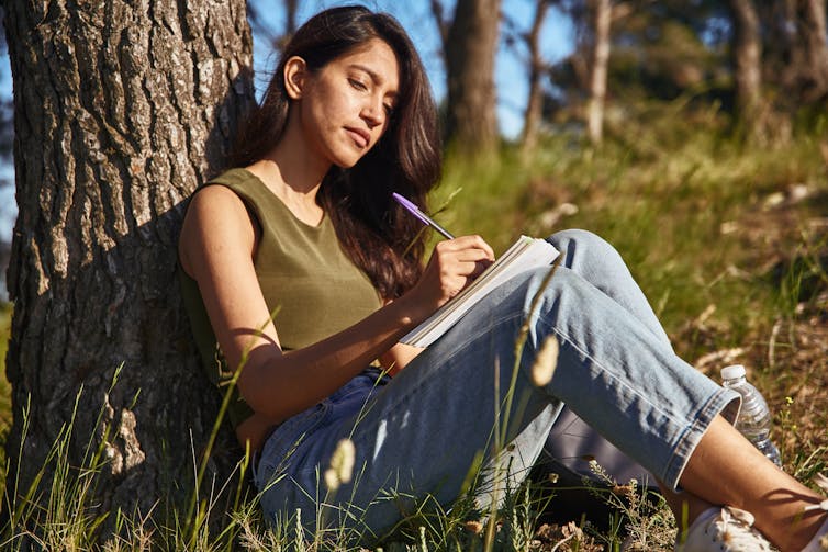 A young woman sitting outdoors at the base of a tree on a sunny day, writing in a journal