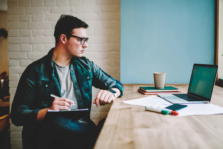 A young man sitting at a counter, looking focused at his laptop and taking notes on a clipboard