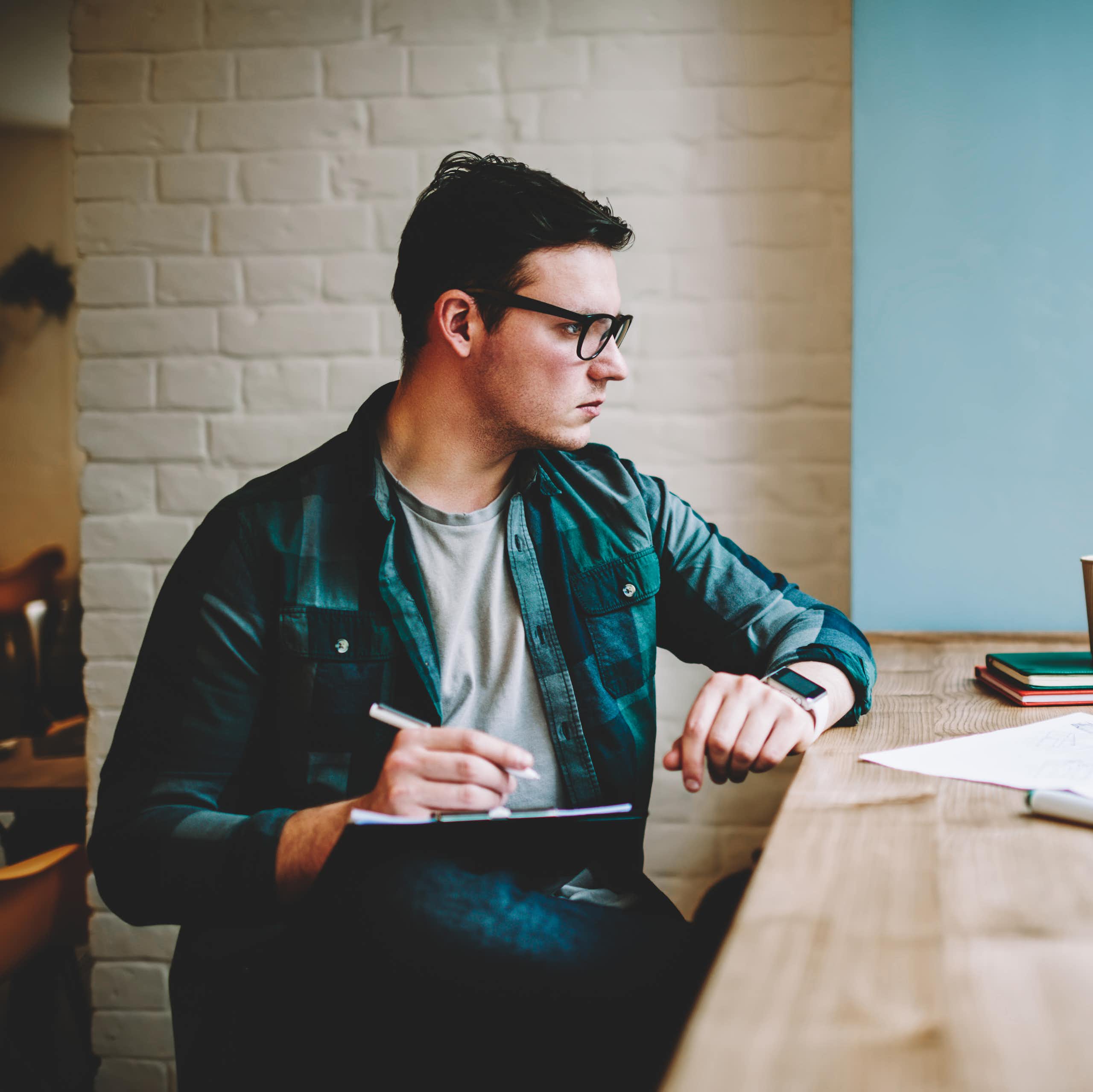 A young man sitting at a counter, looking focused at his laptop and taking notes on a clipboard