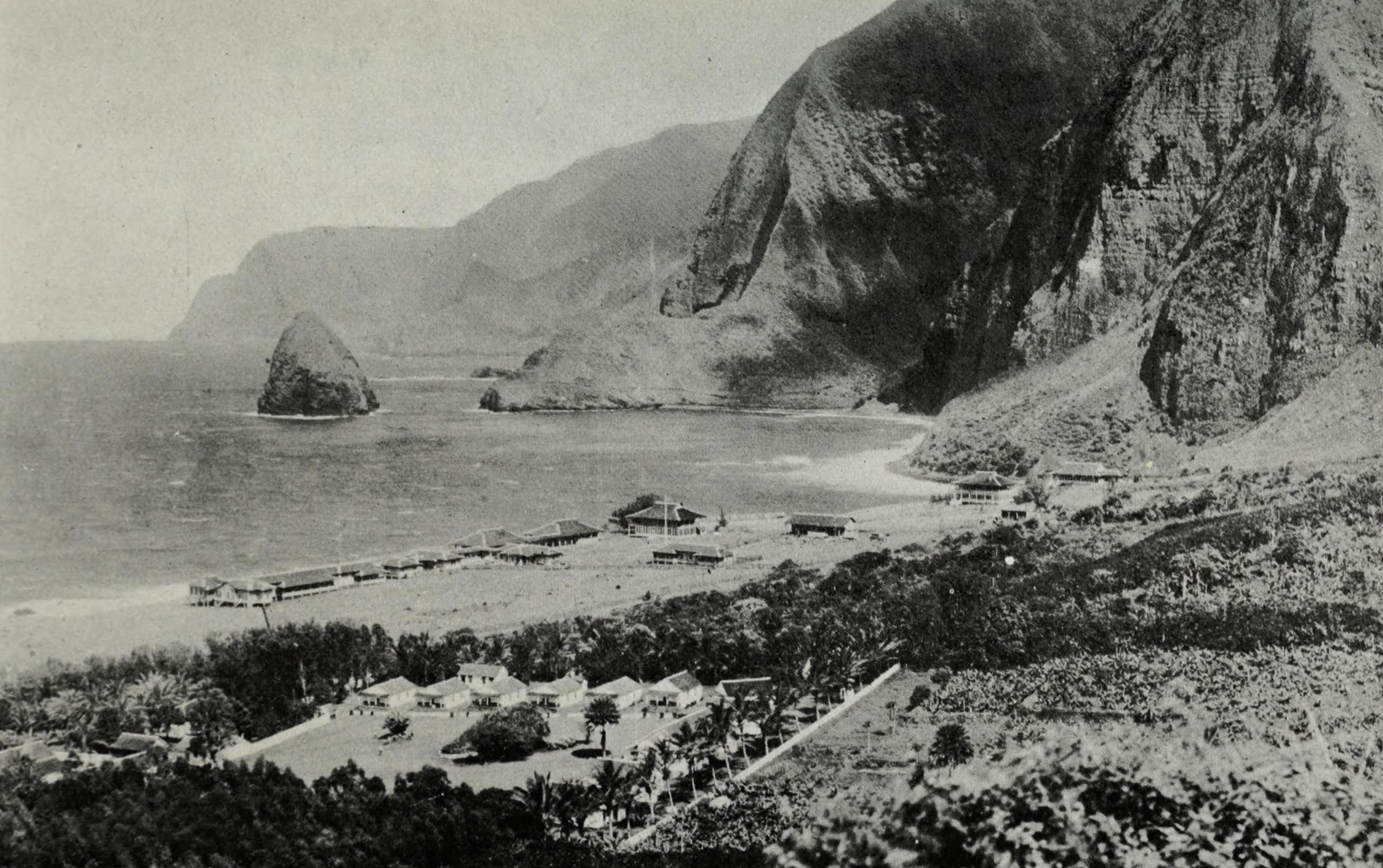 A black and white photo of a small settlement of buildings on a coast, with cliffs visible in the background.