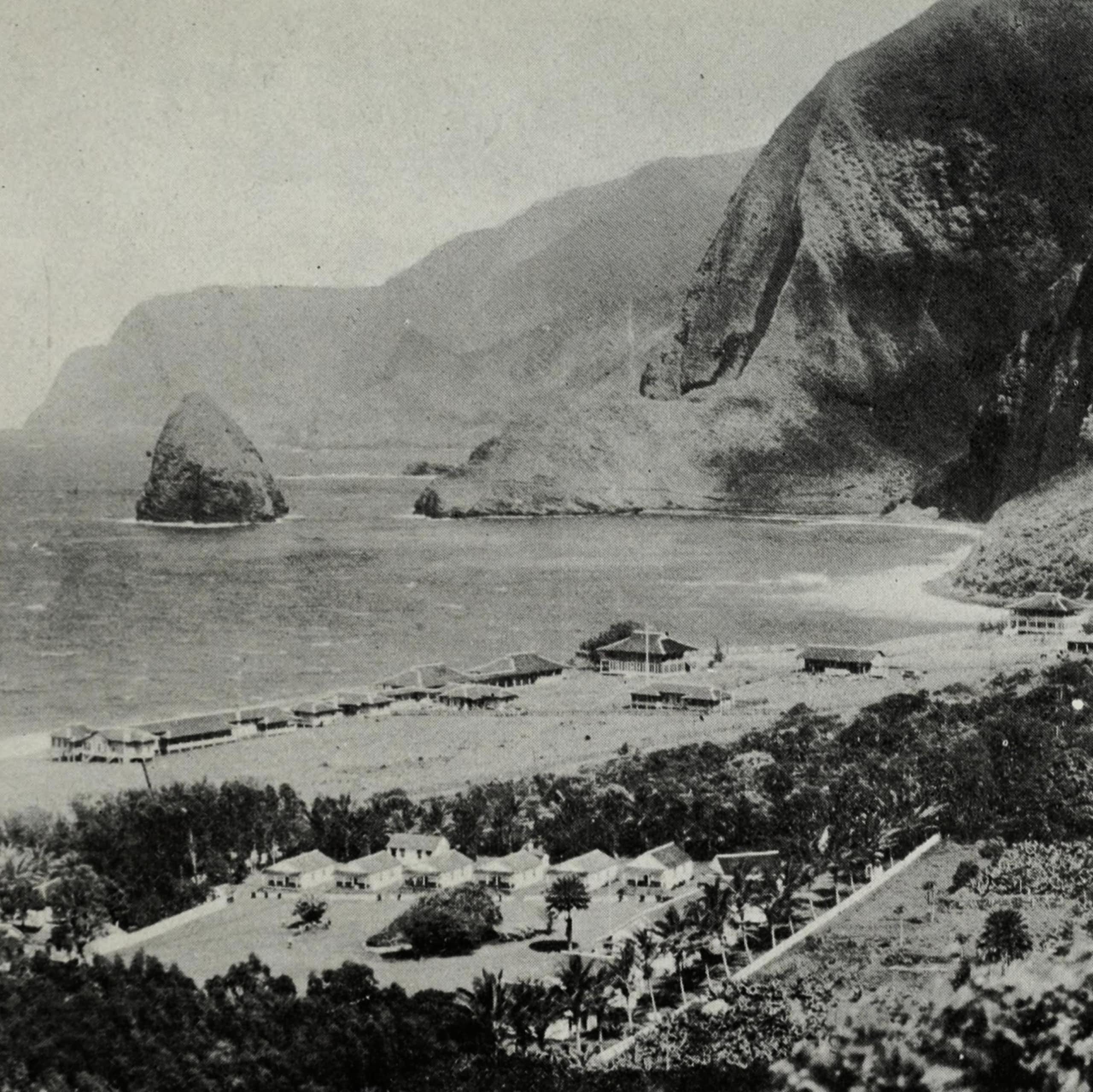 A black and white photo of a small settlement of buildings on a coast, with cliffs visible in the background.