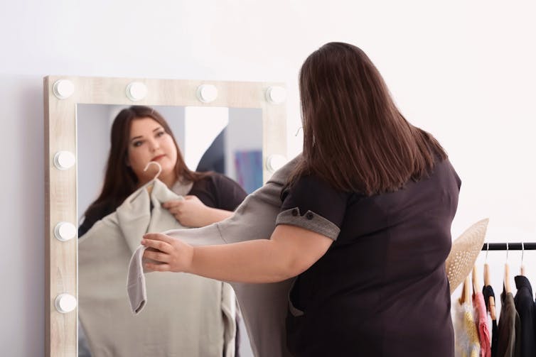 A plus-sized woman in front of a mirror holding a dress up to herself