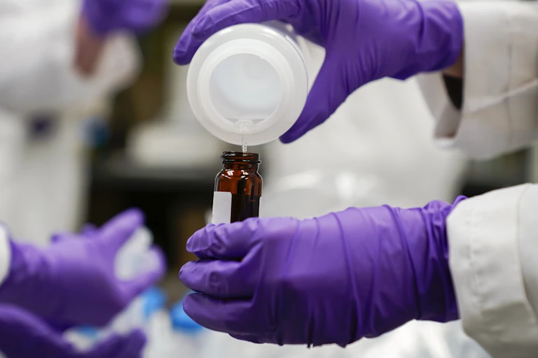 Hands in purple gloves pour a water sample into a bottle for testing.