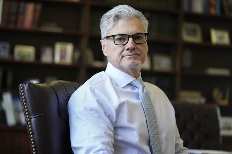 A gray-haired man in a dress shirt and tie, sitting in a study.