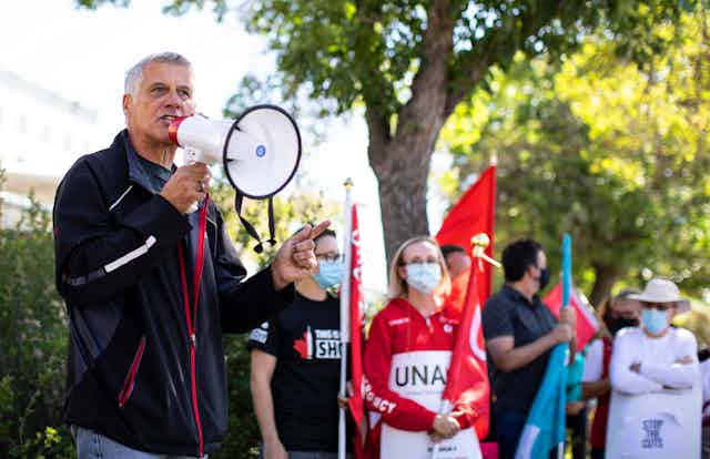 A middle-aged man speaks into a megaphone while a crown of people holding red flags look on