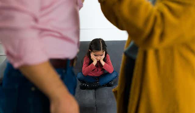 A young girl with her head in her hands sits sadly between two bickering parents