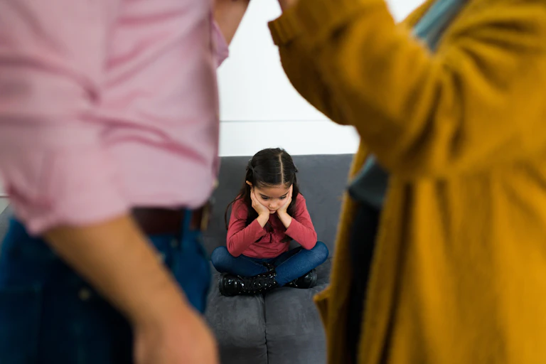 A young girl with her head in her hands sits sadly between two bickering parents