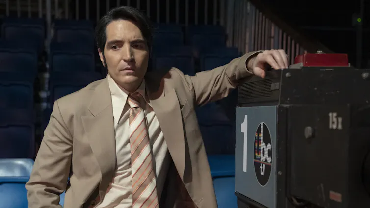 A man in a ’70s-style TV host suit stands in an empty studio, leaning on a camera.