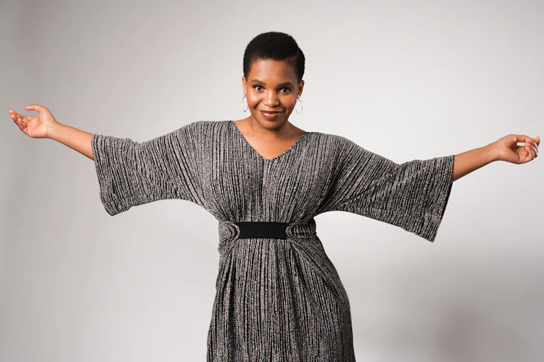 An African woman with short, natural hair smiles directly at the camera and extends her arms, wearing a gown that is pleated and drawn together in the waist.