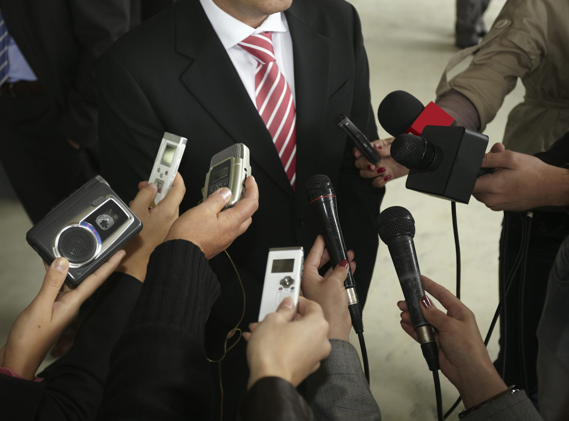 A group of journalists with recording devices listening to a man in a suit make a statement.