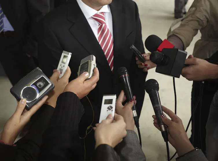 A group of journalists with recording devices listening to a man in a suit make a statement.
