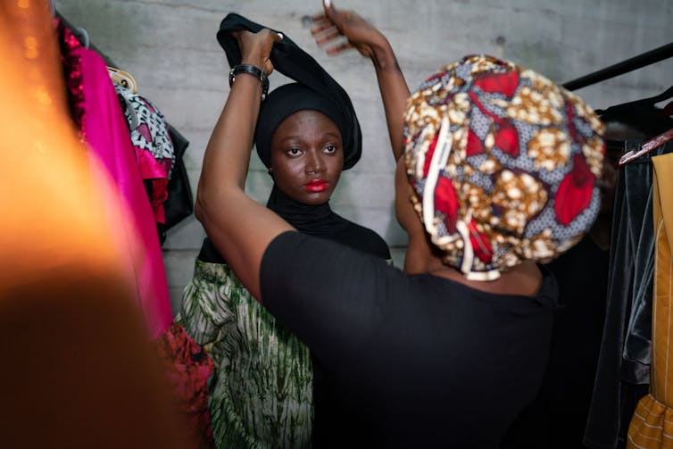 A woman standing next to a railing of clothing has her head wrap tied by another woman, who wears a head wrap in African wax print cloth.