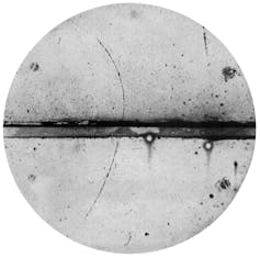 Cliché en noir et blanc d’une chambre à brouillard. Le positron est entré dans la chambre en bas à gauche, a été ralenti par la plaque de plomb (ligne épaisse au milieu du cliché) et sa trajectoire s’est incurvée en haut à gauche