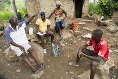 A man in a yellow tshirt, the author of this article, sits on a rock on sandy terrain taking notes as four other men, all fishermen, describe their plight with Lake Kariba's water levels falling