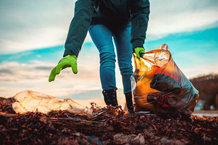 Shot of man wearing jeans, green gloves, leaning forward with plastic bag in left hand, right hand reaching for plastic bottle in foreground among seaweed, beach in distance, blue cloudy sky