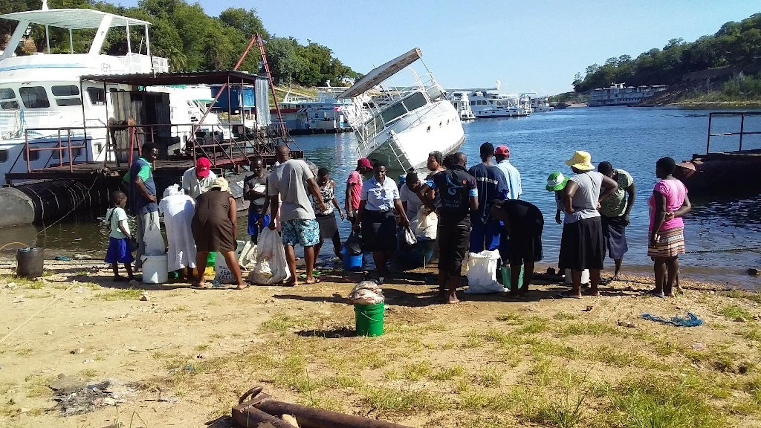 About 18 people stand on the shores of Lake Kariba near small fishing trawlers buying and selling fish