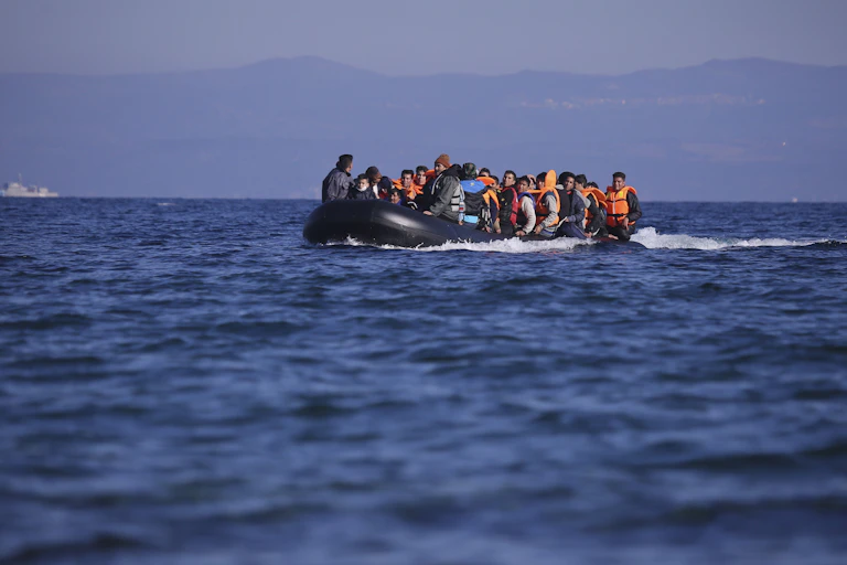 A dinghy full of men in lifejackets on a blue sea in 2015, reaching the Greek island of Lesbos