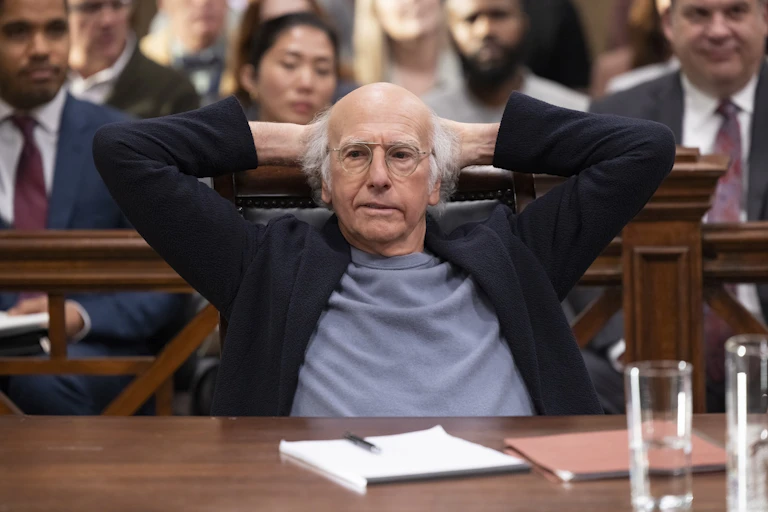 Larry David sits at a desk with his hands behind his head