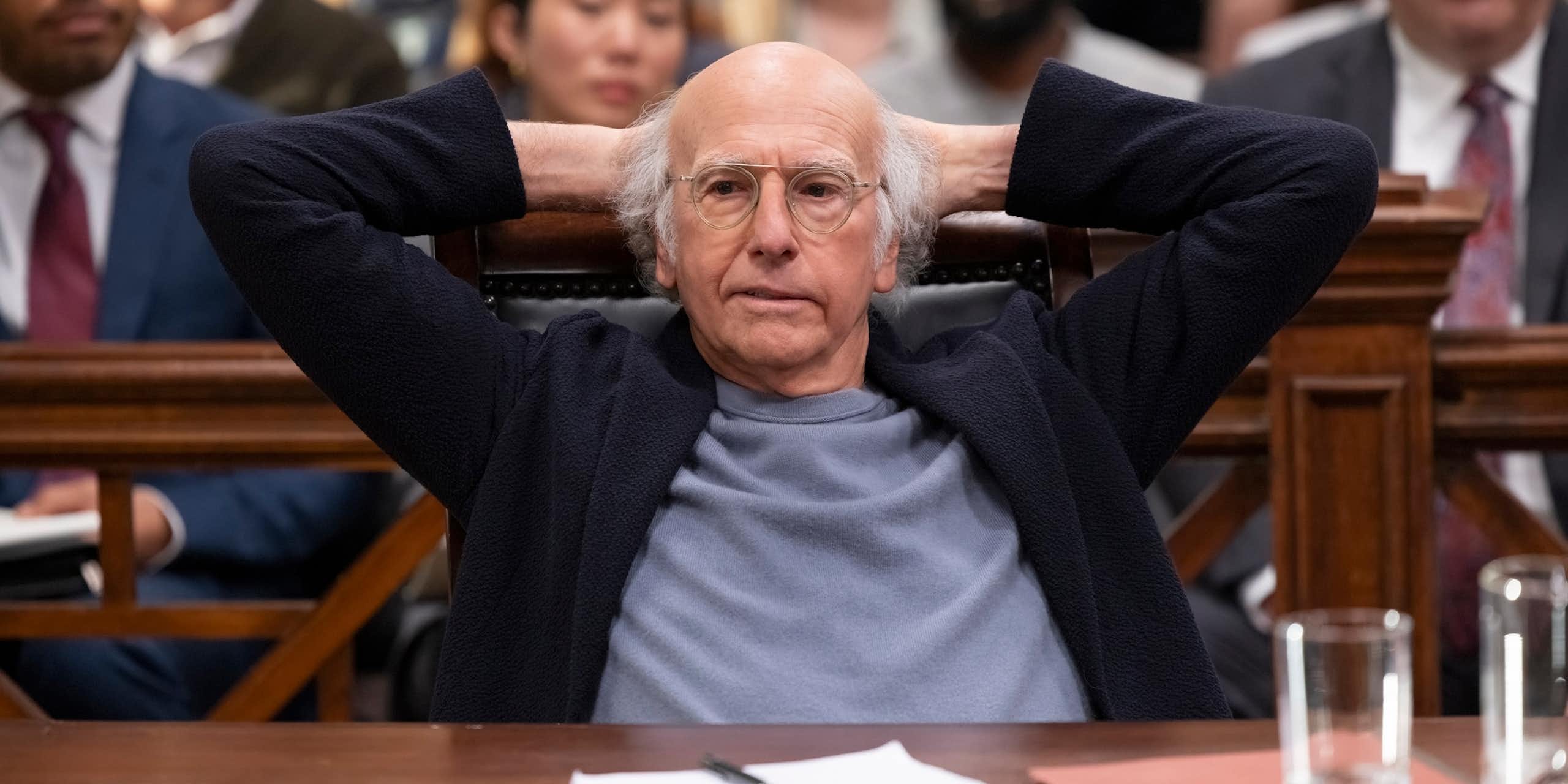 Larry David sits at a desk with his hands behind his head