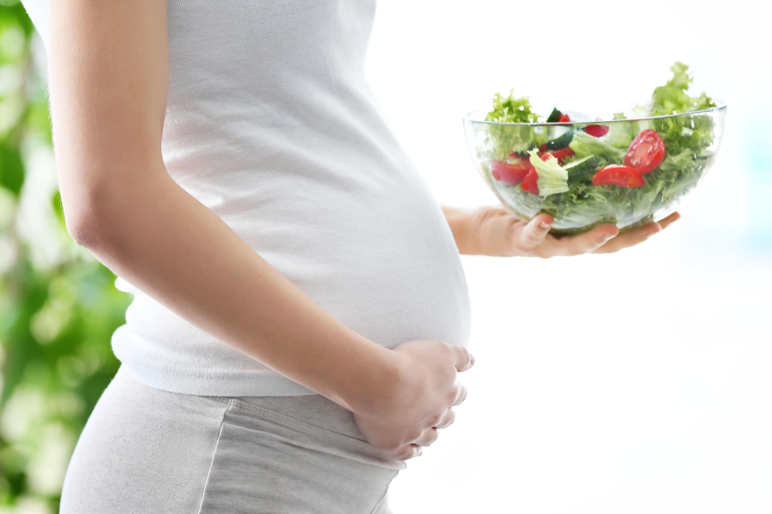 Une femme enceinte de profil tient son ventre d'une main et un bol contenant une salade de crudités de l'autre main.