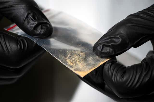 Gloved hands holding small clear packet of brown powder substance