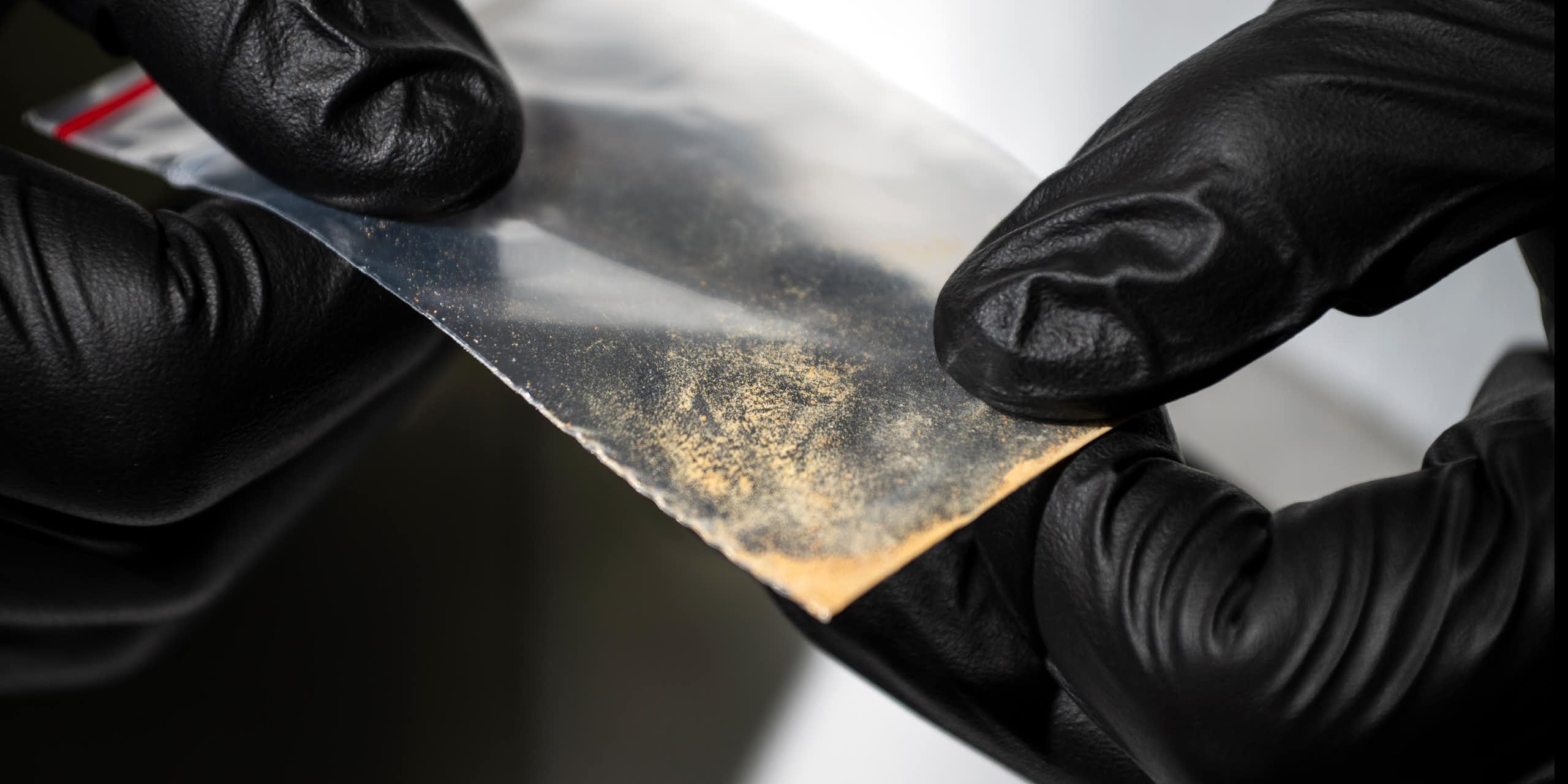 Gloved hands holding small clear packet of brown powder substance